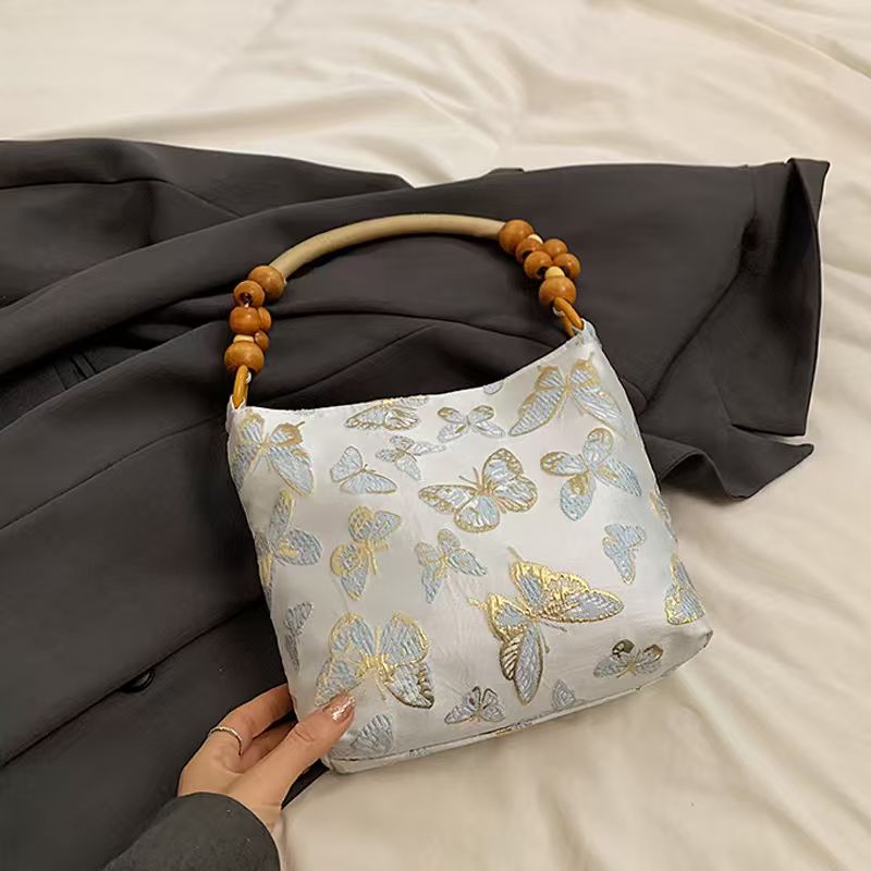 A white shoulder bag with a gold and blue butterfly pattern, featuring a curved wooden handle with brown beads, held by a hand over a dark gray blazer and white bedsheet background.