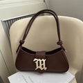 A dark brown leather shoulder bag with a large gold metal 'M' monogram on the front, resting on a white book against a cream-colored chair.