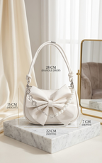 A white handbag with a large bow and pearl trim sits on a marble pedestal in a softly lit interior. Black measurement text overlays show the bag's dimensions: 28 cm handle drop, 15 cm height, 22 cm width, and 7 cm depth.